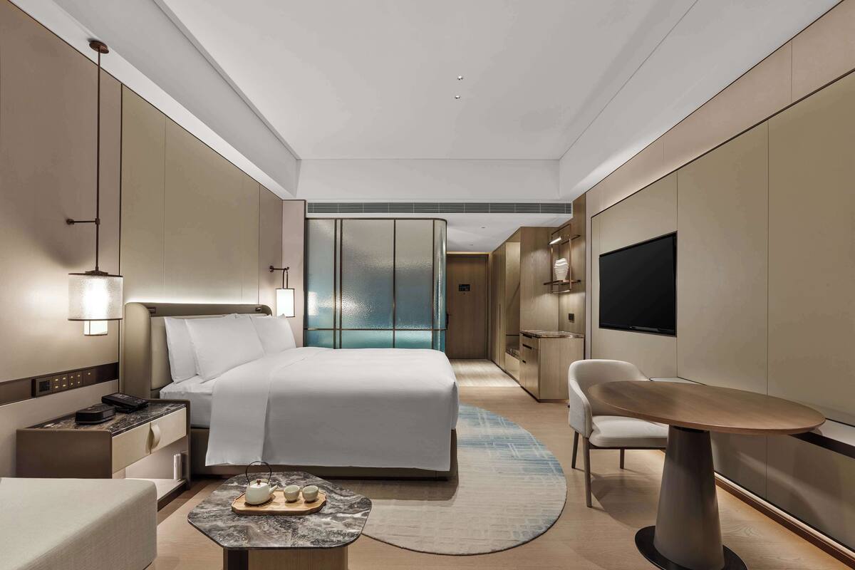 Quarto de hotel moderno com uma cama grande, mesa de cabeceira, televisão e um espaço de estar com mesa e cadeiras.