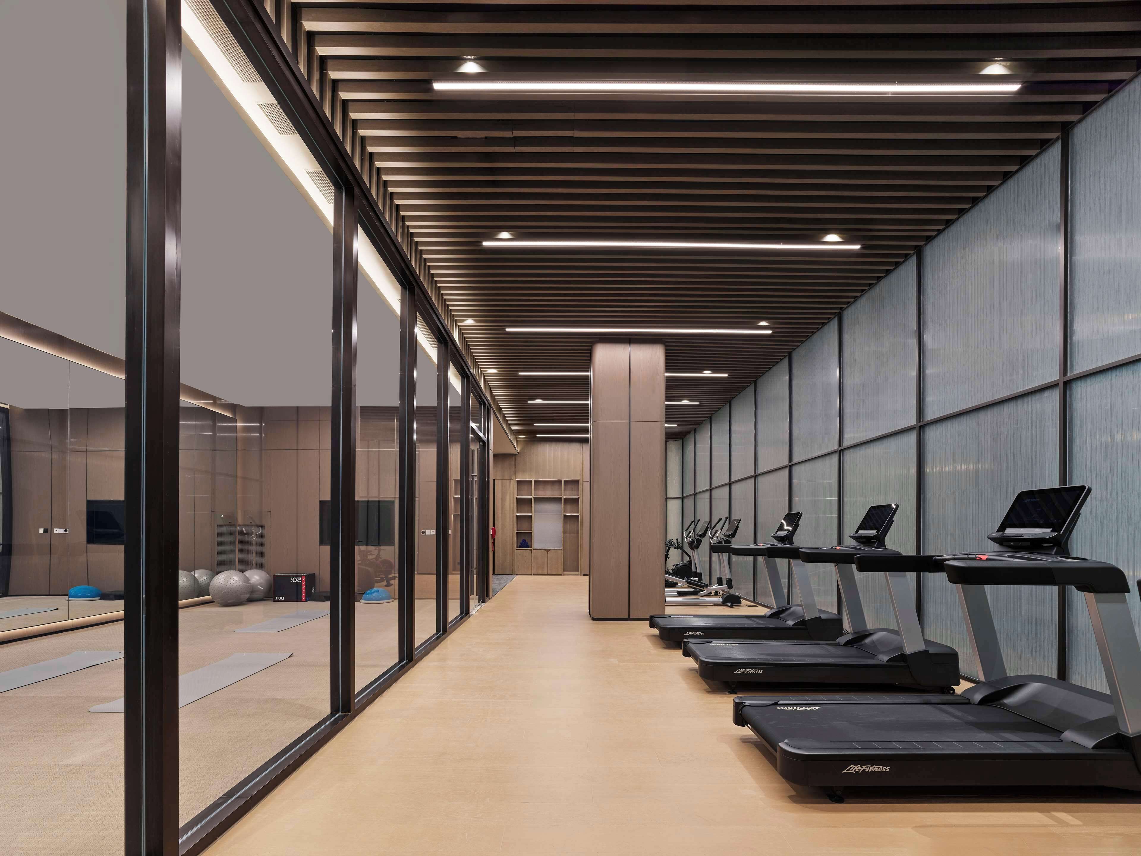 A modern gym with treadmills aligned along a corridor, large windows, and a wooden floor.