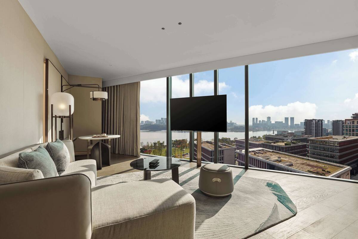 Quarto de hotel moderno com uma grande janela, sofá, mesa de centro, televisão e vista para o horizonte da cidade.