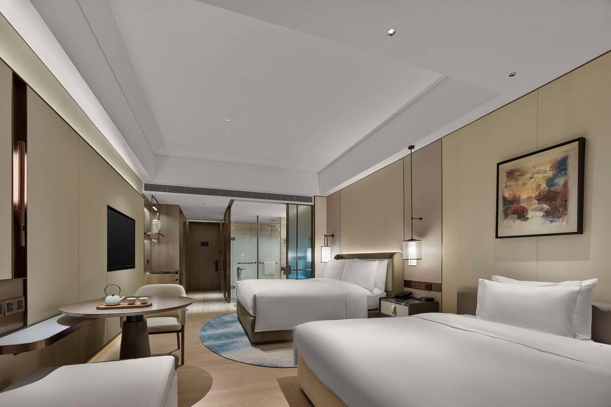 Quarto de hotel com duas camas, uma escrivaninha, uma cadeira, uma televisão e um quadro na parede.