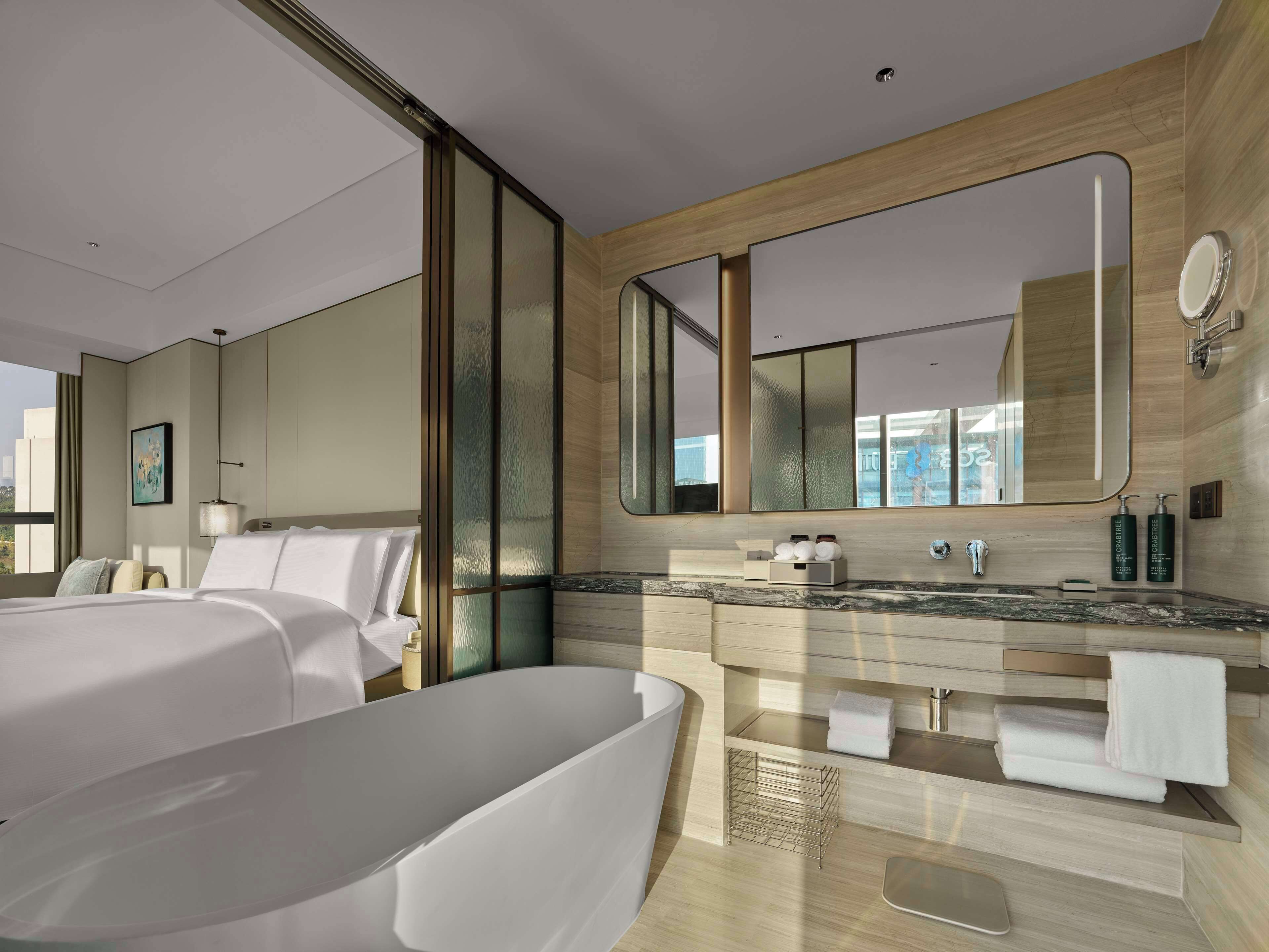 A modern hotel room with a large bed, a separate sitting area, a large mirror, a vanity with a sink, and a view of the city.
