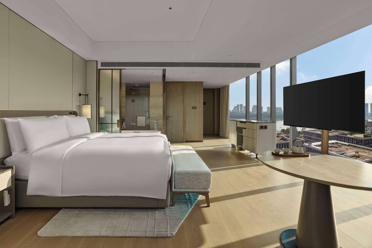 Quarto de hotel moderno com uma cama grande, uma escrivaninha, televisão e vista para a cidade.