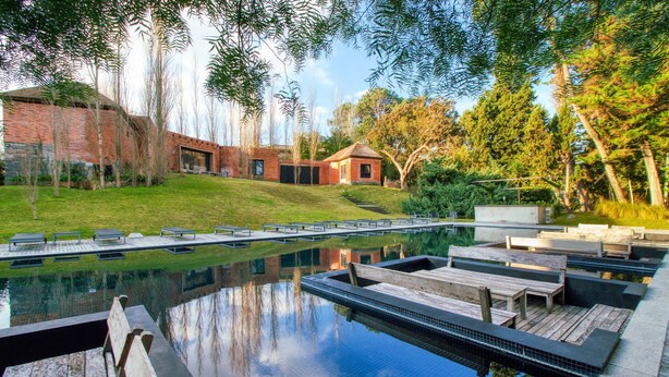 Piscina con mobiliario de terraza de madera, edificio de ladrillo y vegetación circundante.