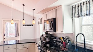 Una cocina moderna con armarios blancos, encimera negra y horno empotrado. Hay luces colgantes, una ventana con cortinas y suelo de madera.