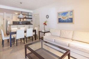 Uma sala moderna com mesa de centro de vidro, sofá branco, mesa de jantar com cadeiras e uma cozinha ao fundo.