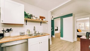 Una cocina moderna con muebles blancos, encimera de madera y lavavajillas integrado. Hay una repisa sobre el fregadero con diversos utensilios de cocina.
