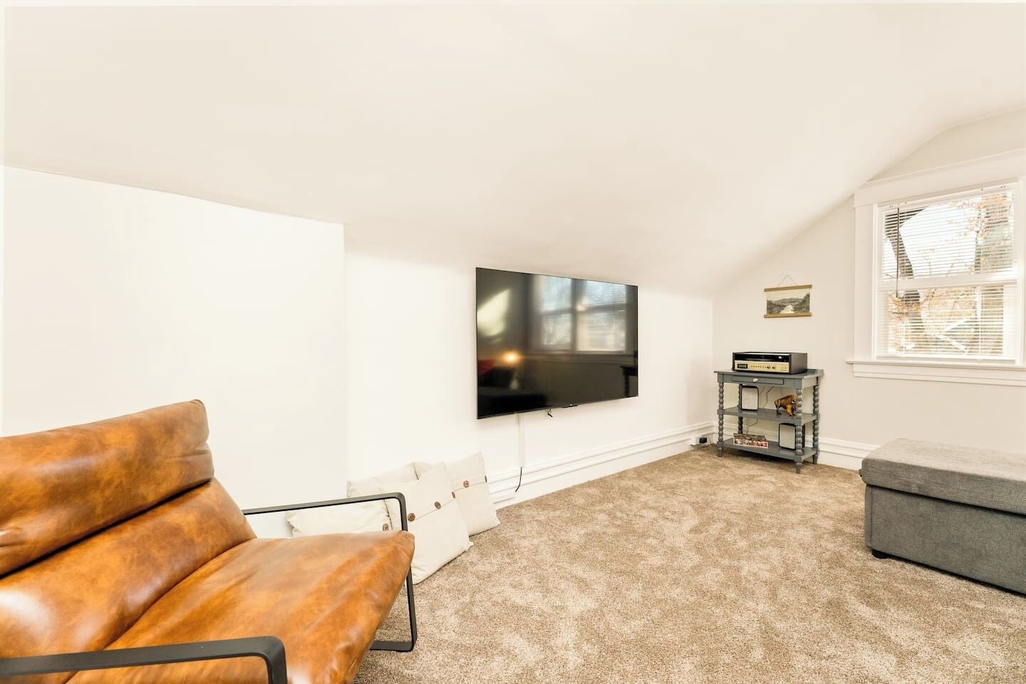 A living room with a leather sofa, a flat-screen TV mounted on the wall, a small table with electronic devices, and a window with blinds.