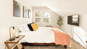 Un dormitorio con cama, mesita de noche, lámpara, ventana y televisor montado en la pared.