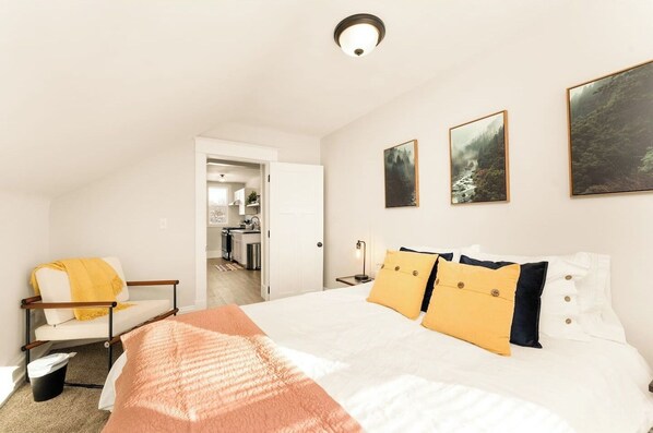 A bedroom with a bed, a chair, a bedside table, and a door leading to a kitchen.