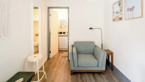 A compact living space with a grey sofa, a small wooden table, a white stool, and a doorway leading to a kitchen area.