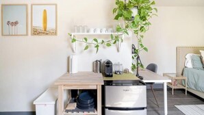 A compact kitchen with a refrigerator, a wooden table, and a hanging plant. There are framed pictures on the wall and a bed in the background.