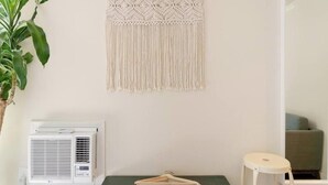 A room with a macrame wall hanging, a small table with a hanger, a potted plant, and a white air conditioning unit.