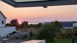Vue depuis une pièce dotée d’une lucarne, donnant sur un paysage côtier avec des maisons et de la végétation, au moment du coucher du soleil.