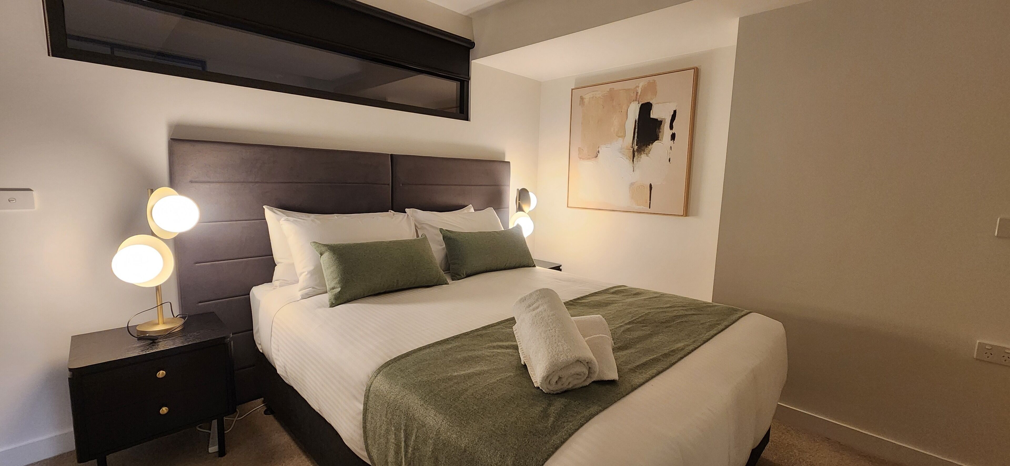 Un lit bien fait, avec une couverture verte et des oreillers blancs, une lampe de chevet et une œuvre d’art accrochée au mur.