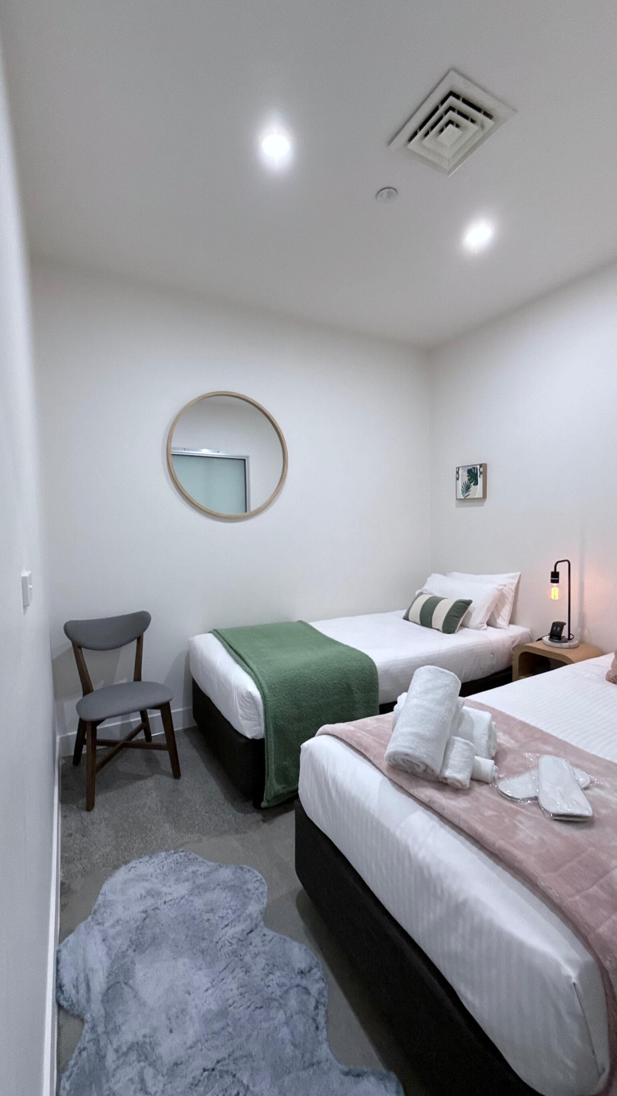Habitación de hotel con dos camas, una silla, una mesita de noche con lámpara, un espejo en la pared y piso alfombrado.