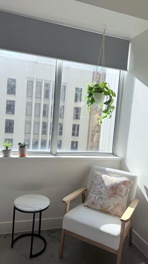 Ein Zimmer mit großem Fenster, einer hängenden Pflanze, einem kleinen Tisch und einem Stuhl mit Blumenkissen.