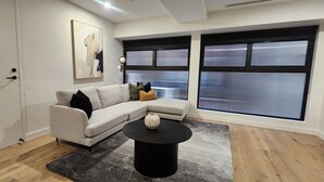 A modern living room with a light-colored sectional sofa, a black coffee table, and a large window with blinds. - Collins St - The Heart Of The City (Melbourne)