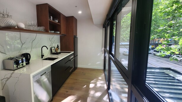 A modern kitchen with a built-in dishwasher, wooden cabinets, and a large window offering a view of the street. - Collins St - The Heart Of The City (Melbourne)
