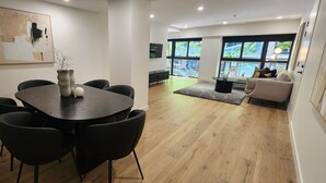 Un intérieur moderne avec une table à manger ronde, des chaises noires, un vase et un canapé, le tout dans une pièce lumineuse dotée d’un plancher en bois et de grandes fenêtres.