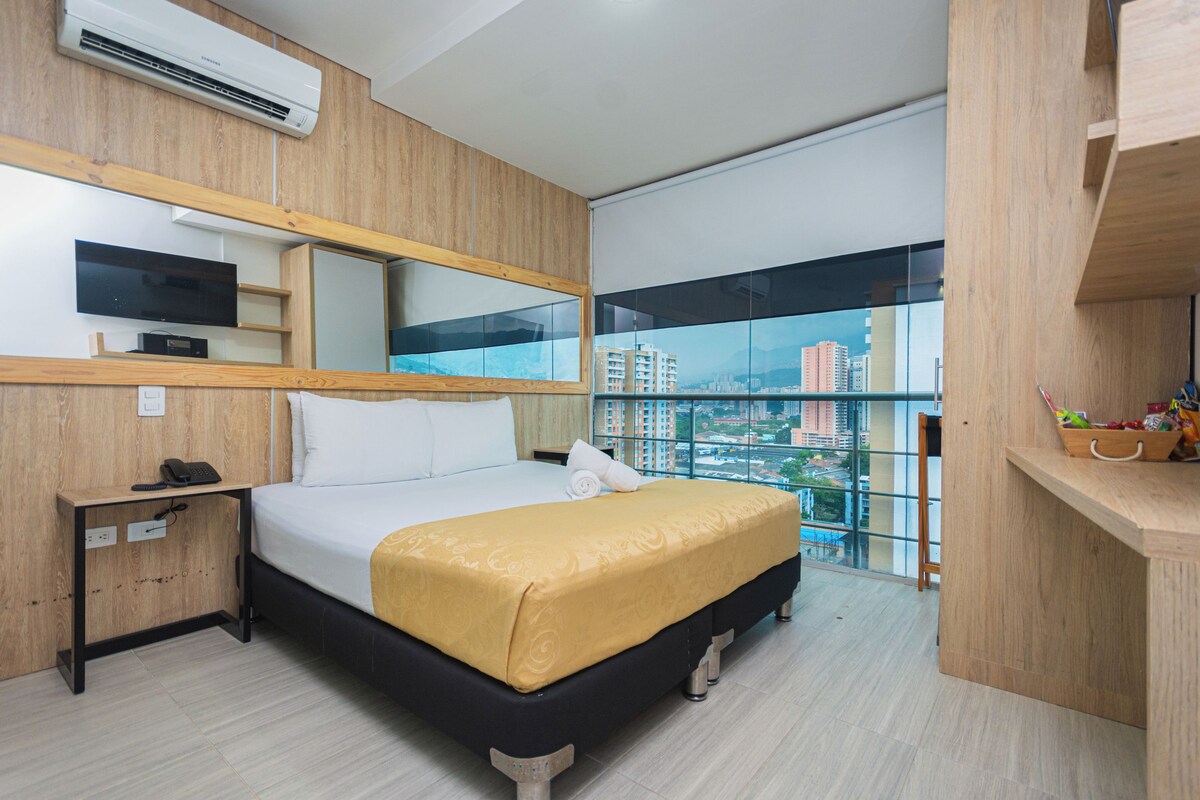 A hotel room with a bed, a bedside table, a mirror, and a window with a city view.