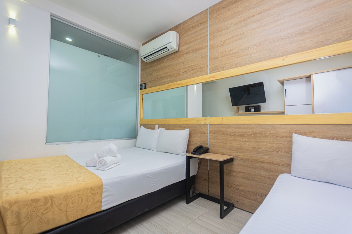 A hotel room with a bed, a bedside table with a phone, a small TV, and an air conditioning unit mounted on the wall.
