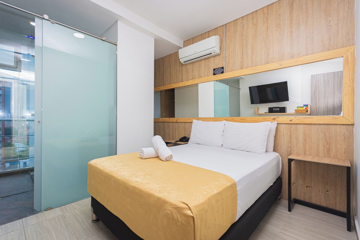 A hotel room with a bed, a bedside table, a TV, and an air conditioning unit.