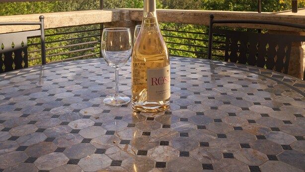 Una mesa con una botella de vino rosado y una copa sobre un piso de baldosas hexagonales, con vistas a un viñedo bajo un cielo despejado.