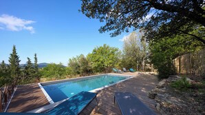 Una terraza de madera con piscina, sillas de descanso y vistas a árboles y montañas.