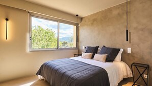 Un dormitorio con un ventanal grande, una cama con ropa de cama oscura, una mesita de noche y una lámpara colgante.