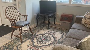 A living room with a wooden chair, a sofa, a television on a stand, a patterned rug, and a window with a view of the outside. - Tiny Cozy Home in Missouri (Bakersfield)
