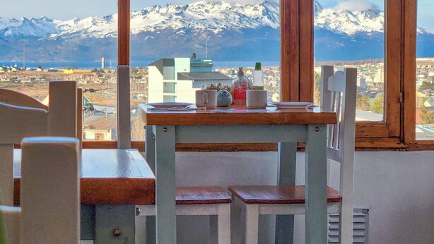 Een kamer met een tafel en stoelen, een raam met uitzicht op besneeuwde bergen en een stadsbeeld.