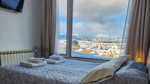 Een hotelkamer met een bed, een nachtkastje met een lamp, een aan de muur bevestigde televisie en een raam met uitzicht op de stad.