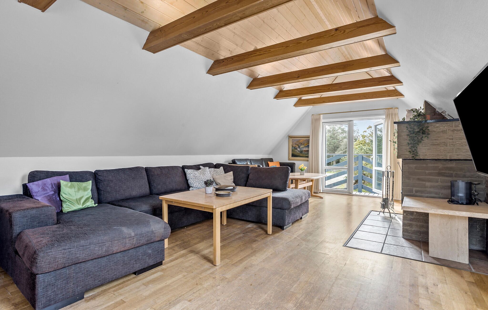 A modern loft with exposed wooden beams, a grey sectional sofa, a wooden coffee table, and a view of the outdoors through a large sliding door.