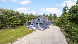 A blue cabin with a gravel driveway, surrounded by trees and a grassy area with a playground. - Stunning home in Vestervig with sauna (Vestervig)