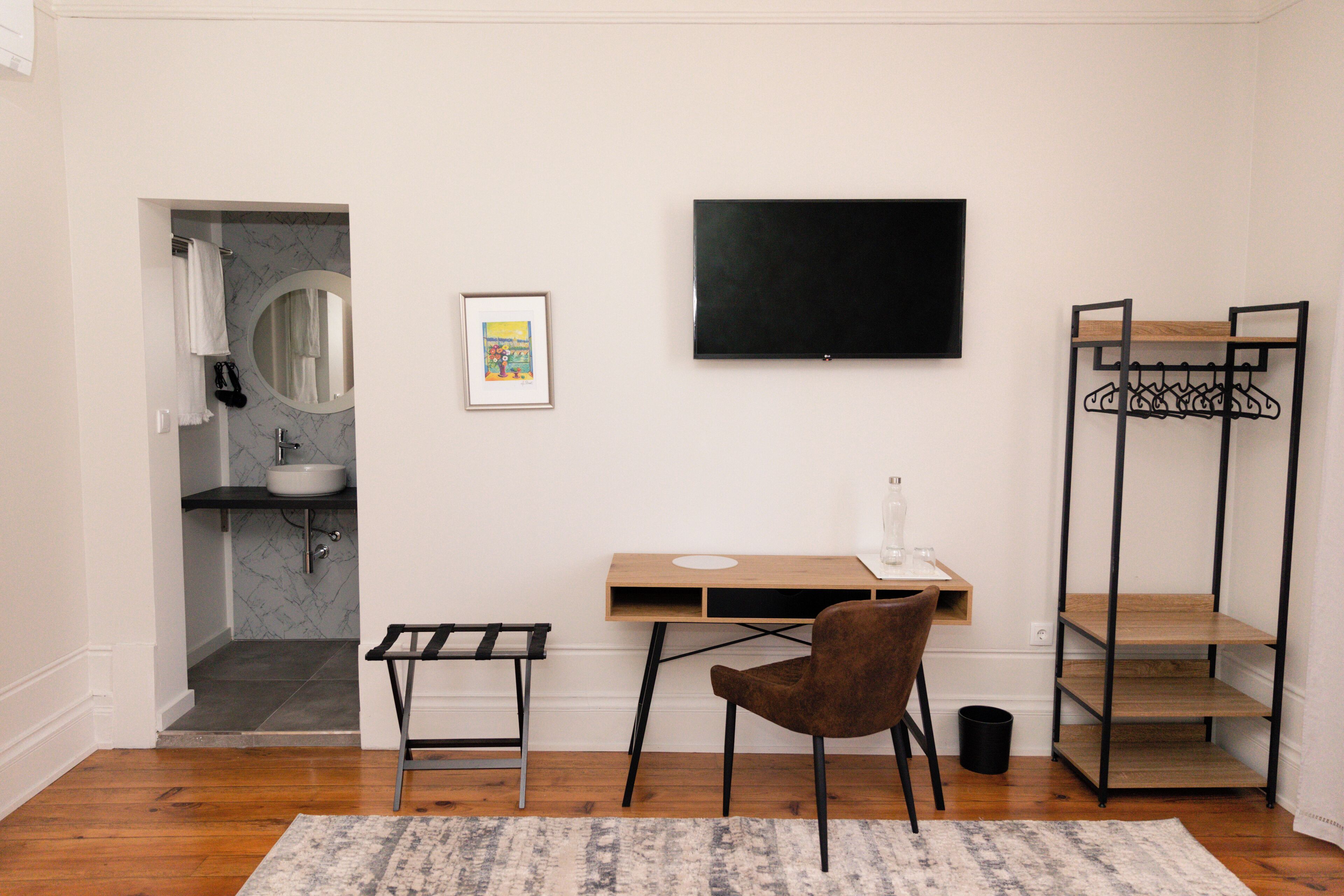 Une petite pièce moderne, équipée d’un bureau en bois, d’une chaise, d’un téléviseur, d’un miroir, d’un lavabo et d’un porte-manteau.