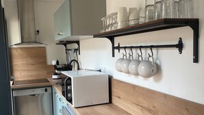 A modern kitchen with a dishwasher, oven, and a wooden countertop. There are shelves with glassware and a hanging rack for cups. - Studio Vichy (Vichy)