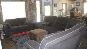 A living room with a stone wall, a grey sofa, a brown ottoman, and a dining area with a table and chairs.