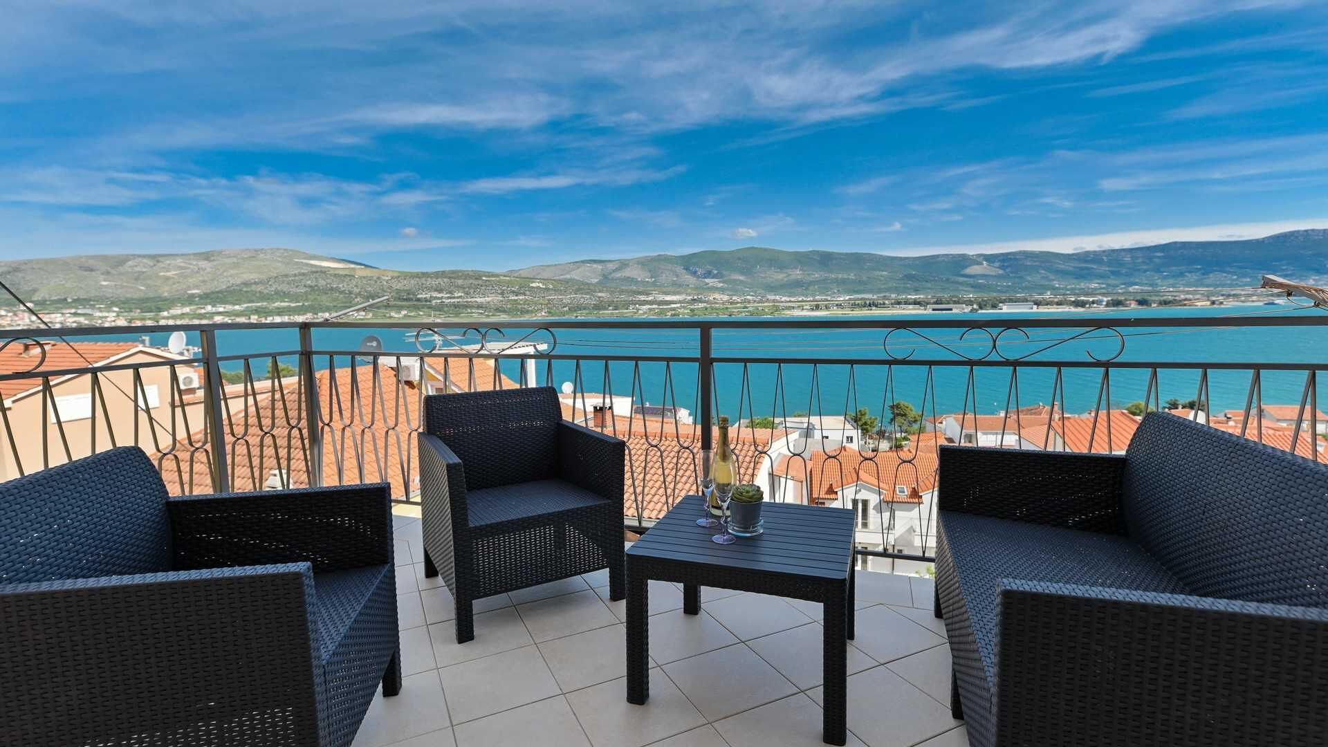 A balcony with wicker furniture and a table, overlooking a coastal view with buildings and hills.