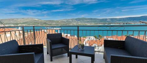 A balcony with wicker furniture and a table, overlooking a coastal view with buildings and hills.