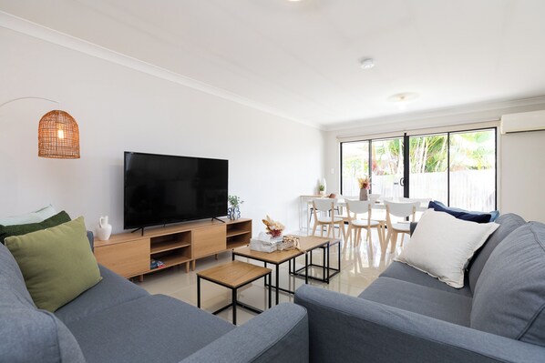 Un salon moderne doté d’un téléviseur à écran plat, de canapés gris, d’une table basse en bois et d’un coin repas avec des chaises et une table blanches.