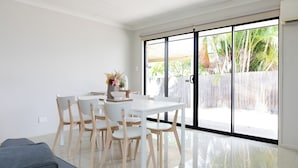 Un espace repas moderne avec une table blanche et des chaises en bois, un vase rempli de fleurs et une vue, grâce à de grandes portes-fenêtres coulissantes, sur un espace extérieur clos.