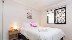 Een netjes opgemaakt bed met witte lakens en twee kussens, een nachtkastje met een lamp, en een raam met een hor.