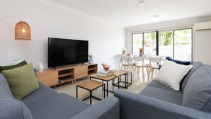 A modern living room with a flat-screen TV, grey sofas, a wooden coffee table, and a dining area with white chairs and a table. - Private Acacia Ridge 1BR | Kitchen & Access | 6BS1 (Acacia Ridge)