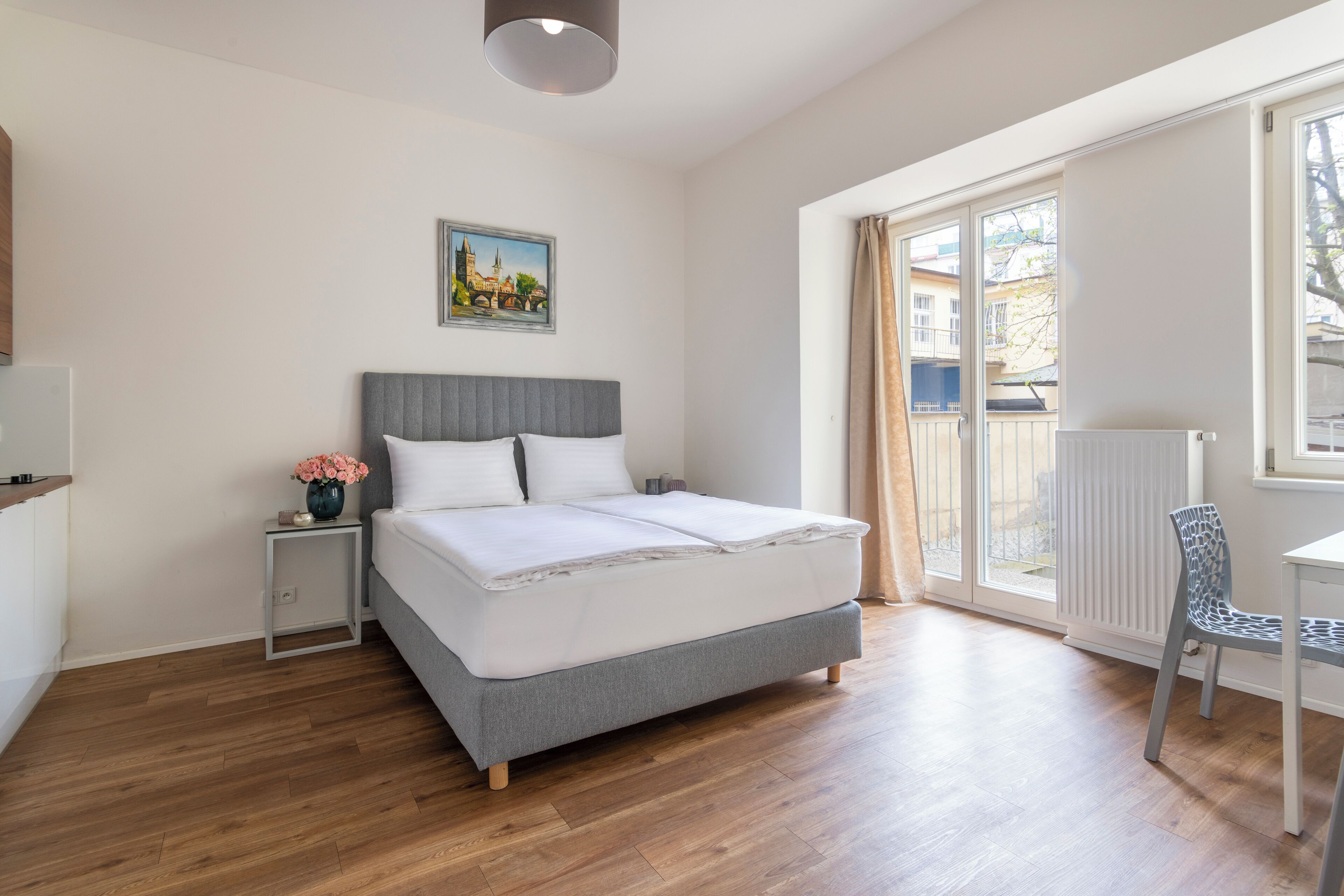 A modern bedroom with a large bed, a bedside table with a vase of flowers, a chair, and a window with a view of buildings.
