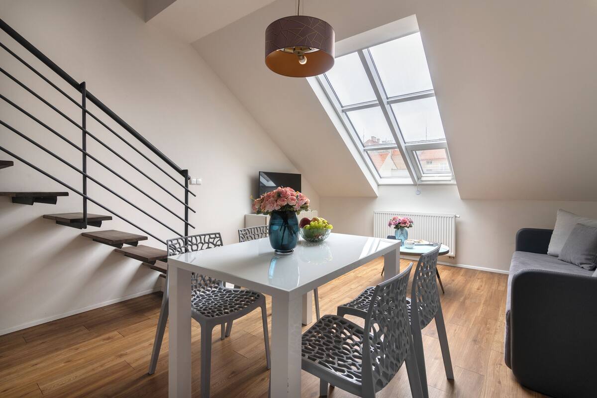 Un appartement moderne de style « loft » avec une table à manger blanche, des chaises noires, un vase bleu avec des fleurs, un canapé gris et un escalier avec des rampes noires.