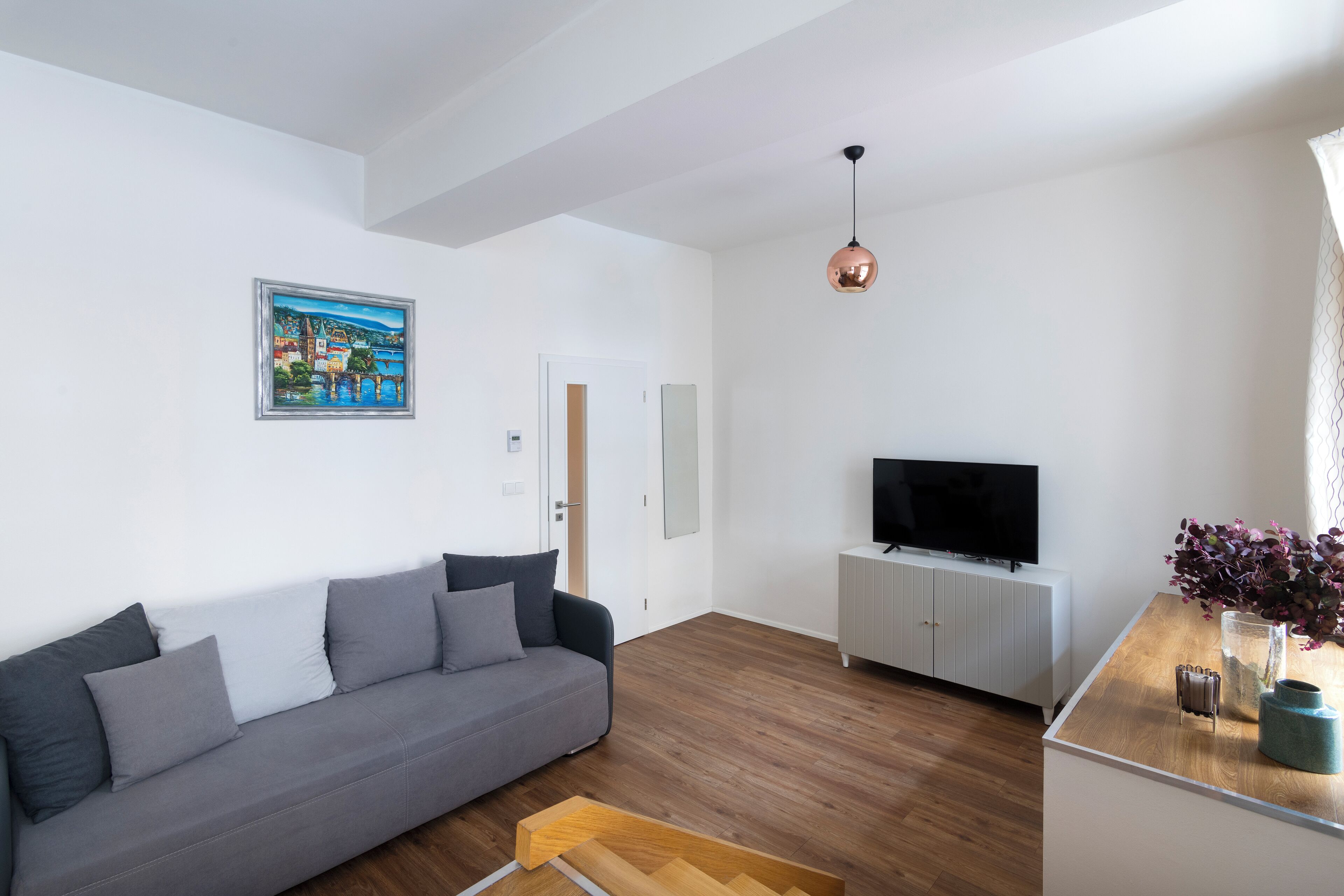 Una sala de estar moderna con un sofá gris, una mesa de centro de madera, un televisor sobre un mueble blanco y un cuadro en la pared.