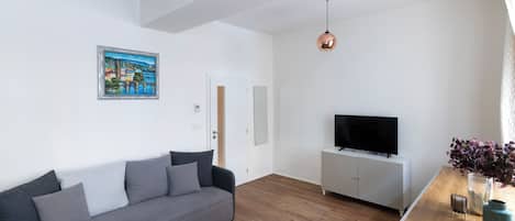 Una sala de estar moderna con un sofá gris, una mesa de centro de madera, un televisor sobre un mueble blanco y un cuadro en la pared.