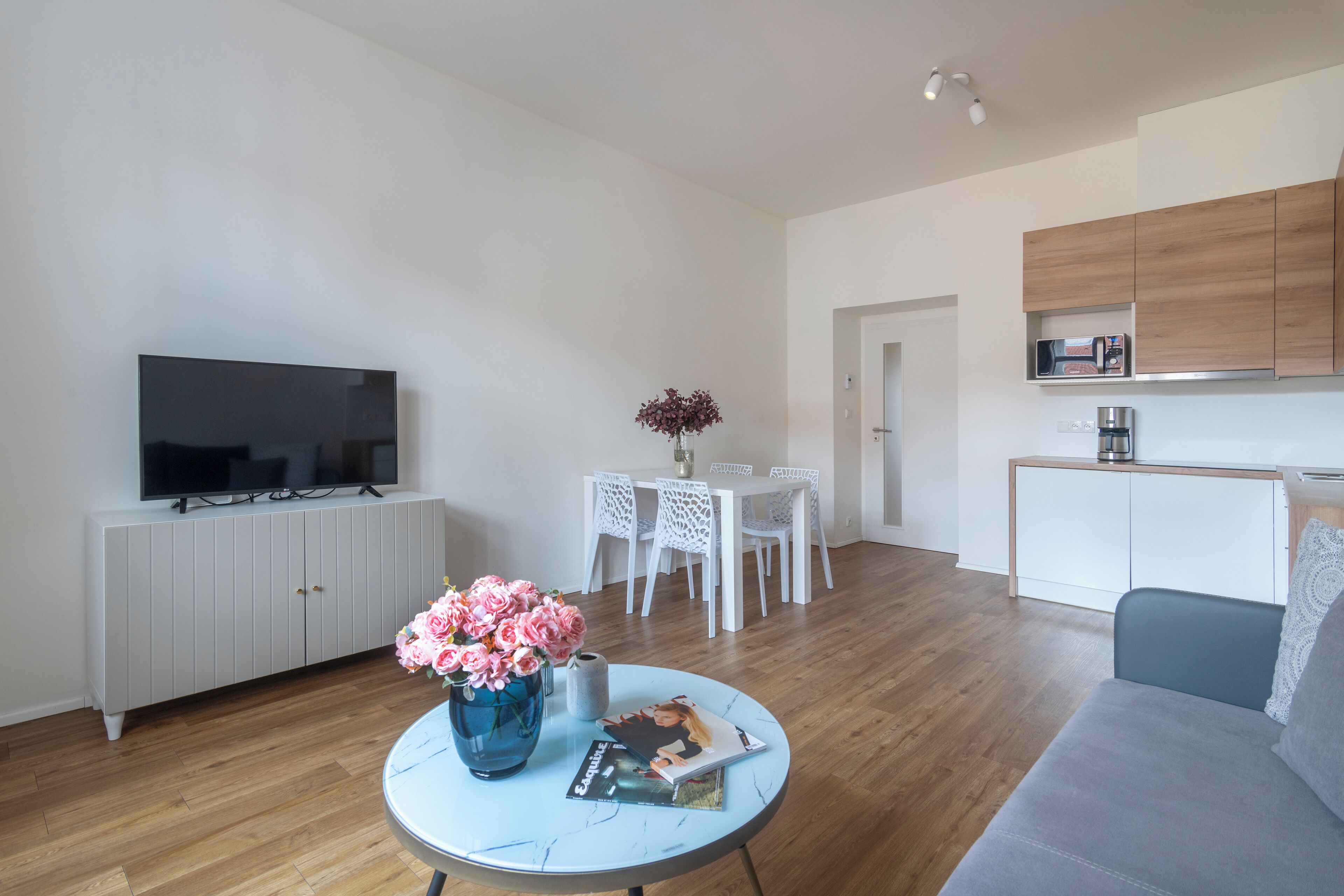 A modern living room with a flat-screen TV, a dining table with chairs, a kitchen area with cabinets, and a sofa.
