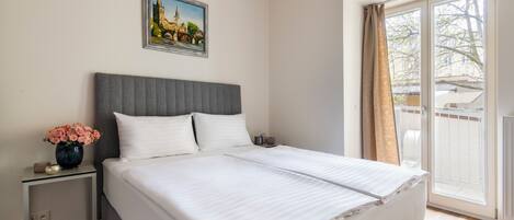 Una cama bien hecha con ropa de cama blanca, un cabecero tapizado en gris y una mesita de noche con un jarrón de flores.