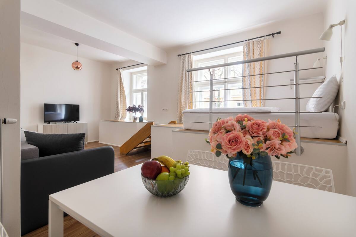 Un salon moderne avec un canapé blanc, une table à manger agrémentée d’un panier de fruits et d’un vase de fleurs, un téléviseur et une fenêtre avec des rideaux.